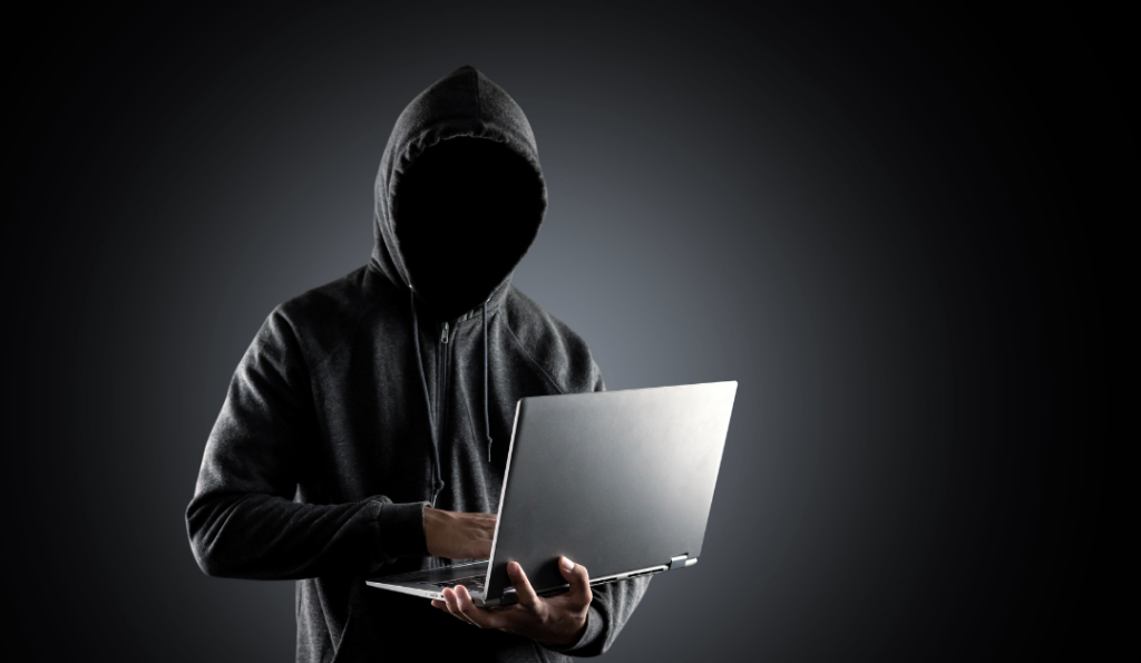 Hacker standing in a hoodie with a laptop in his hands.