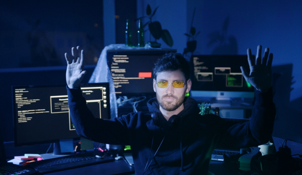 Guy siting with hands in the air and computers behind him.